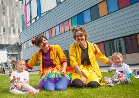 Photo image of two adults dressed as clowns beside two children sitting on grass (Glasgow Children's Hospital Charity Image)