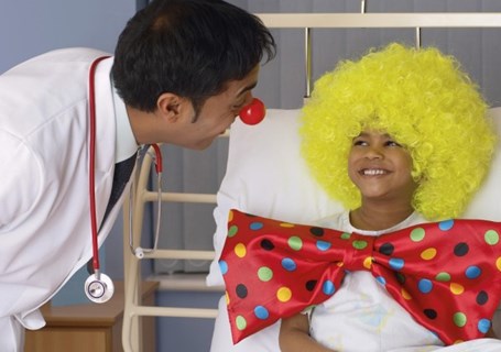 Photo image of an adult dressed as a clown talking to a child in a hospital bed also wearing a yellow clown wig and large bow tie (Glasgow Children's Hospital Charity Image)