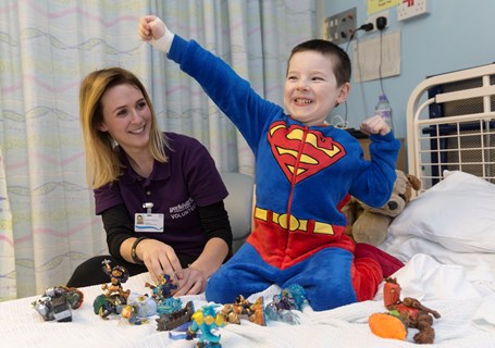 Photo image of a child dressed up as superman sitting on a hospital bed beside a hospital worker (Glasgow Children's Hospital Charity Image)