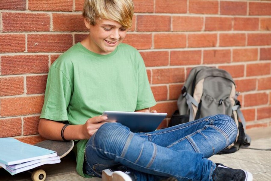 Photo image of a teenager sitting on the ground leaning against a brick wall looking at a tablet (Shutterstock image)
