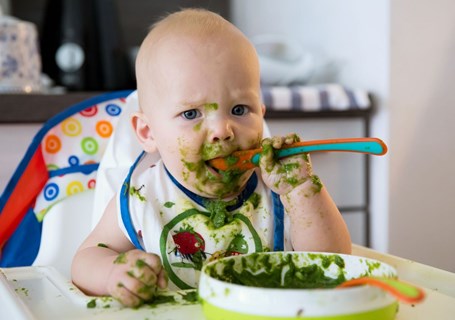 Photo image of a baby sitting in a highchair getting messy by feeding themselves with a spoon (Shutterstock image)