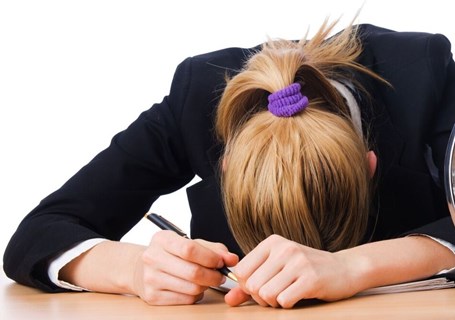 Photo image of a teenager sitting at a desk with a large clock and a large pile of paper.  The teenager has a pen in one hand and the head is resting on the desk (Shutterstock image)